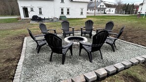 A fire pit surrounded by six Adirondack chairs in a gravel area with a white house in the background. - Spacious, family friendly 5-bedroom home in Pittsburg village with trail access. (Pittsburg)