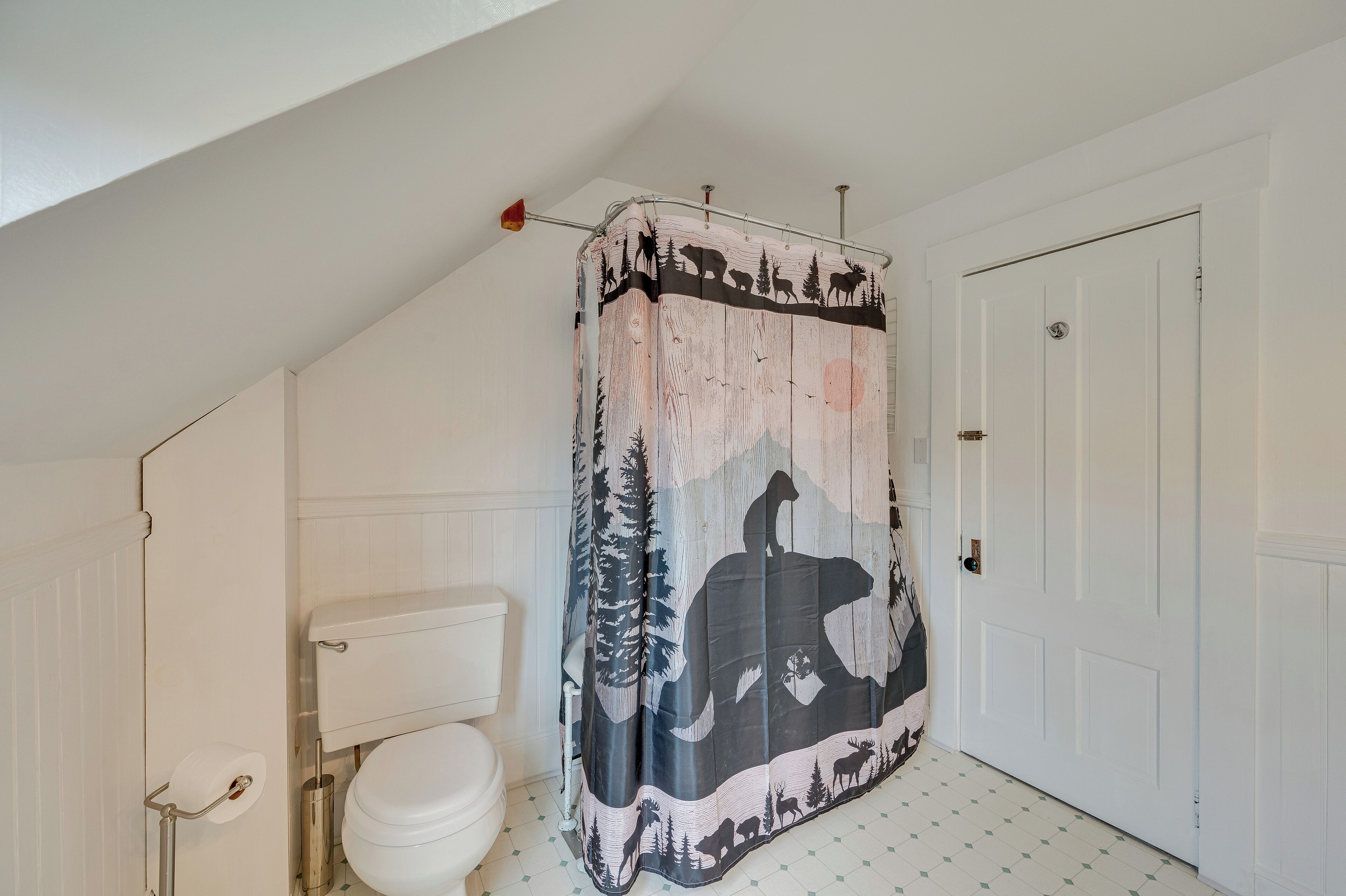 Ein Badezimmer mit einer weißen Toilette, einem Duschvorhang mit Wald- und Tiermotiv sowie einer weißen Tür.