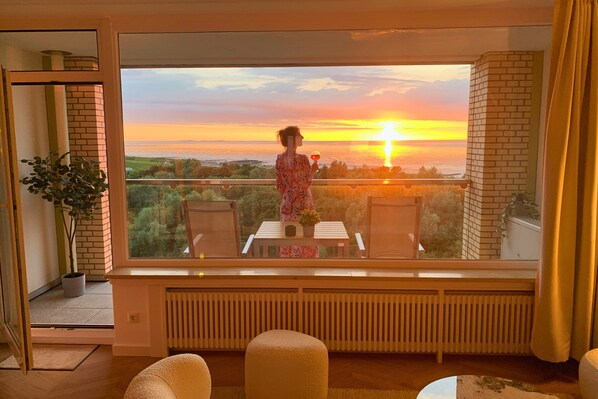 Un balcon avec vue sur le coucher de soleil sur la mer, une table avec des chaises et une plante en pot.