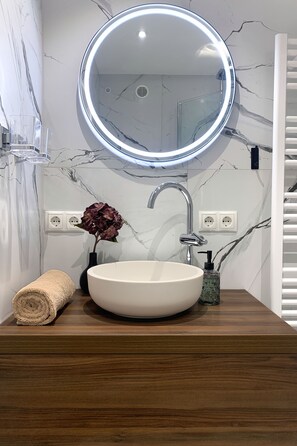 Une salle de bain avec un mur en marbre, un miroir rond doté d’un éclairage à LED, un lavabo blanc, un meuble-lavabo en bois, une serviette et un vase avec des fleurs.