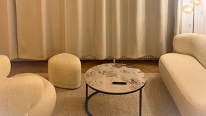 Ein Wohnzimmer mit einem beigen Sofa, einem runden Couchtisch und einem großen Fenster mit beigen Vorhängen.