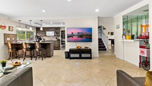 A spacious living area with a kitchen, dining area, and a large TV mounted on the wall. - Fun-Filled Family Vacay Home! 15 min to Disney! (Fullerton)