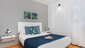 A modern bedroom with a white bed, blue bedding, and tropical-themed wall art. - VILLA DE GRACIA APARTMENT (Barcelona)