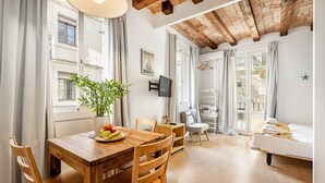 Ein Essbereich mit Holztisch, Stühlen und einer Topfpflanze. Ein Fernseher ist an der Wand befestigt und es gibt ein Fenster mit Vorhängen.