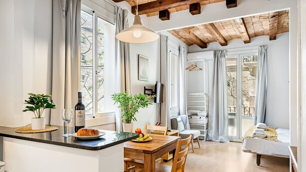 Eine moderne Küche mit einer Holzinsel, einem Esstisch und Stühlen. Auf der Arbeitsplatte stehen eine Weinflasche und Gläser. Der Raum verfügt über große Fenster mit Vorhängen, ein Bett mit weißer Bettdecke und eine Holzbalkendecke.