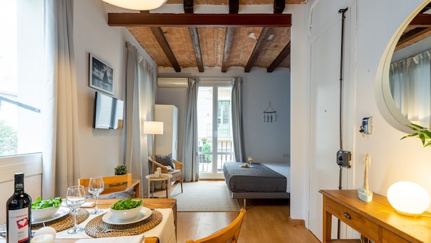 A cozy room with a wooden dining table set for two, a bed with a grey mattress, a small table with a lamp, and a large mirror on the wall. - BARCELONETA BEACH STUDIO 22 (Barcelona)