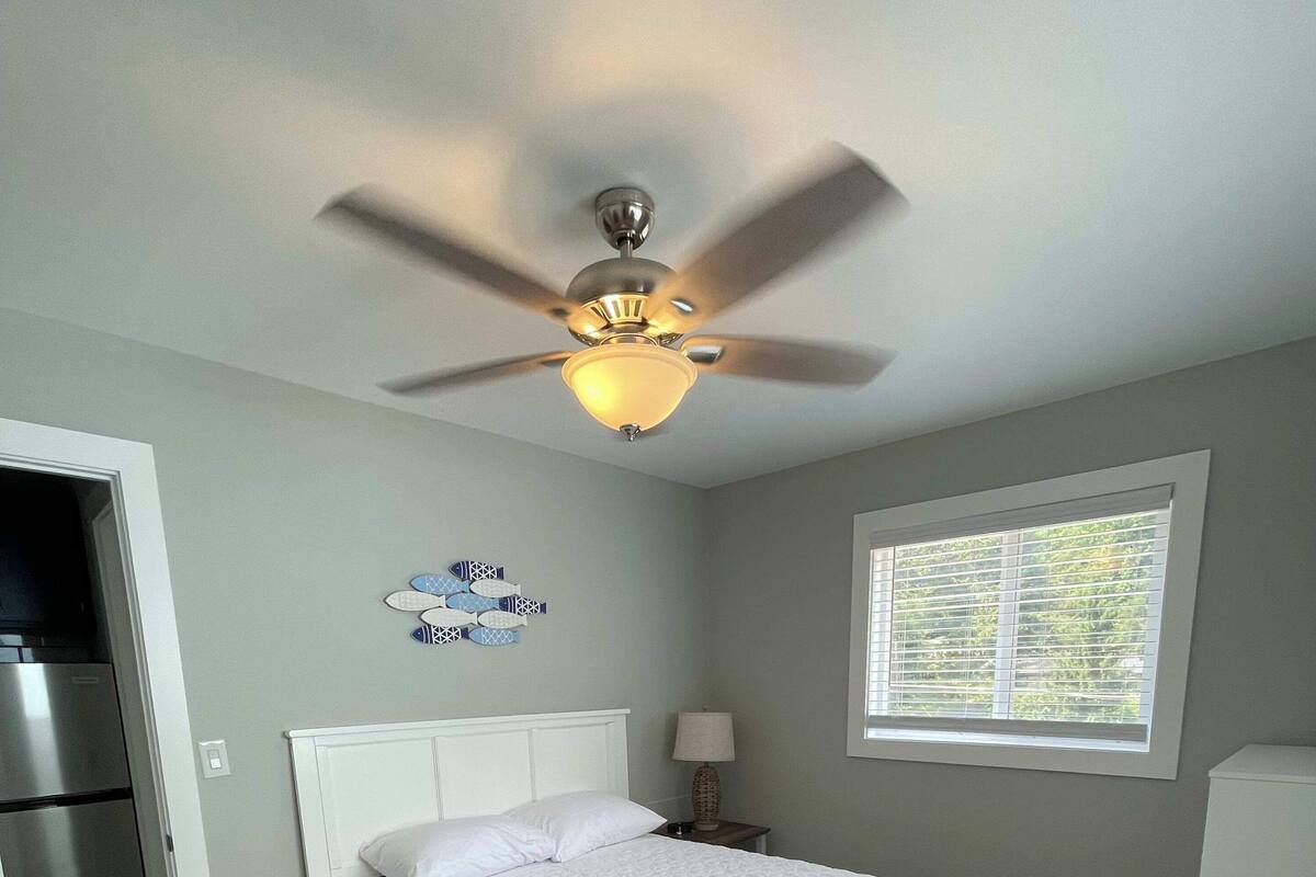 Une chambre à coucher comprenant un lit, un ventilateur de plafond, une fenêtre avec des stores et un réfrigérateur.