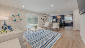 A modern living room with a white sofa, a coffee table, and a striped rug. There is a dining area with chairs and a small table. The kitchen is visible in the background with a window and a hanging light fixture.