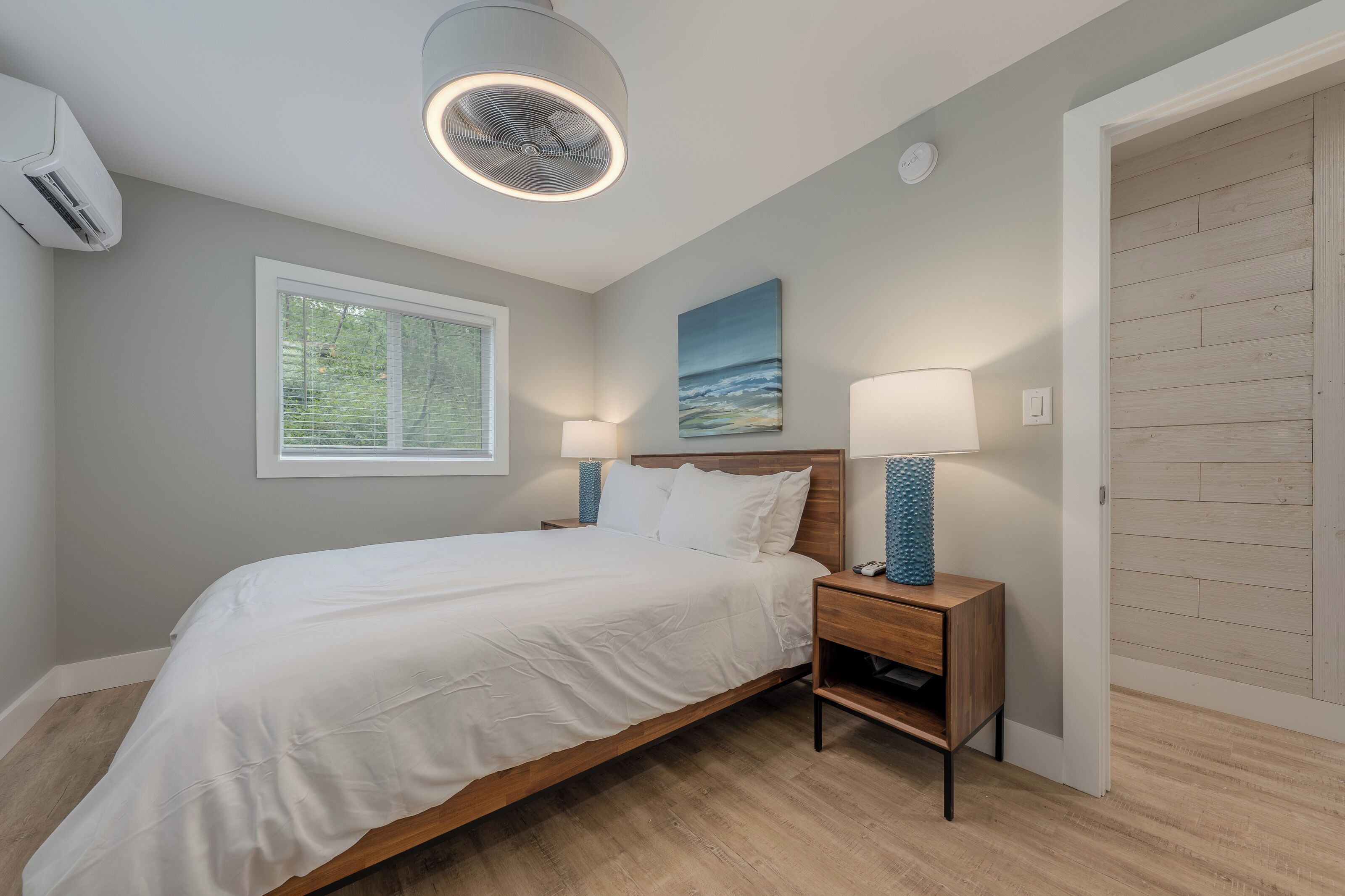 A bedroom with a bed, bedside lamps, a ceiling fan, a window, and a wooden door.