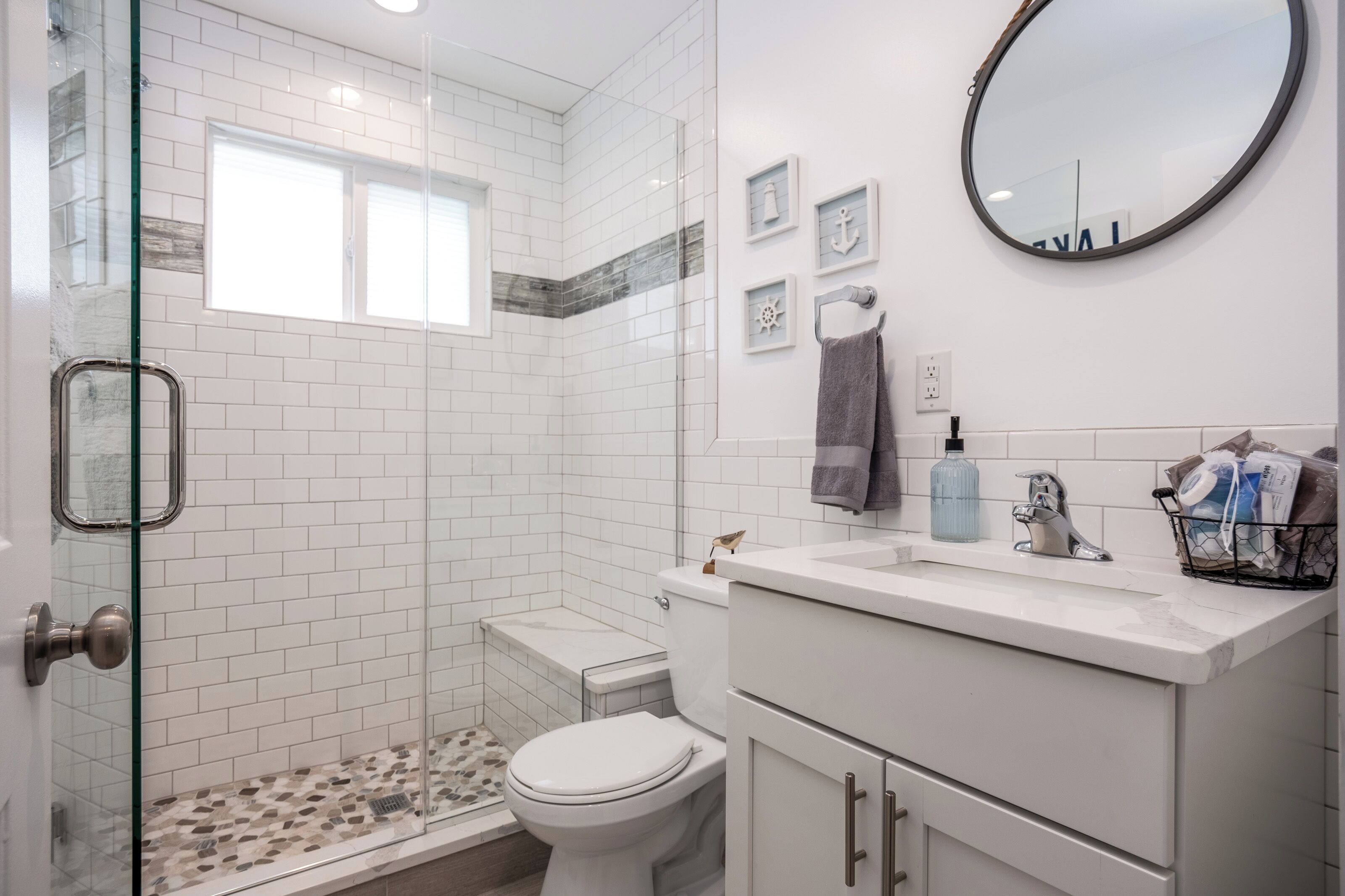 Un baño con cabina de ducha de vidrio, paredes con azulejos blancos, inodoro, lavabo con toallero y espejo.
