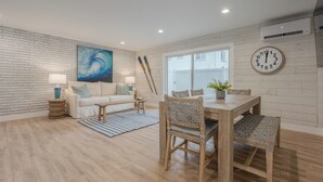 A modern living room with a brick wall, a large abstract painting, a wooden dining table with wicker chairs, a white sofa, and a blue-striped rug.