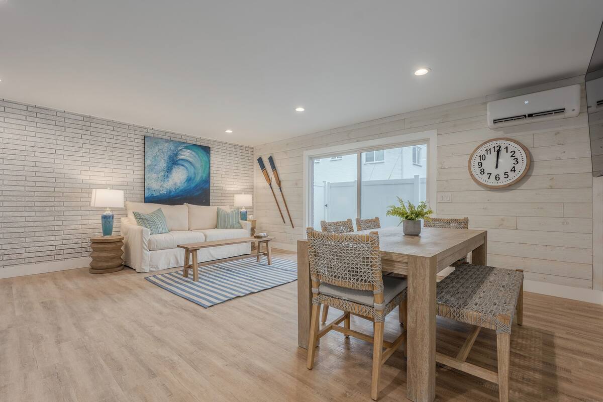 Un salon moderne avec un mur en briques, une grande peinture abstraite, une table à manger en bois avec des chaises en osier, un canapé blanc et un tapis à rayures bleues.