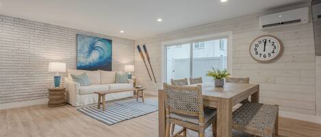 A modern living room with a brick wall, a large abstract painting, a wooden dining table with wicker chairs, a white sofa, and a blue-striped rug.
