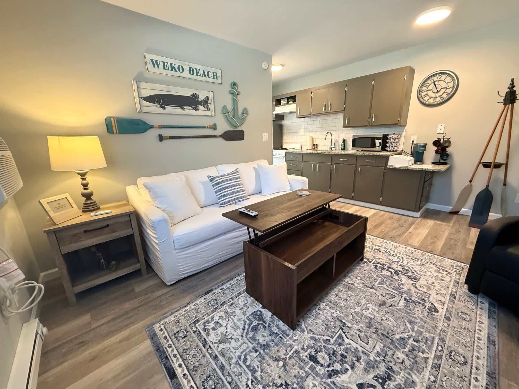 Un salon avec un canapé blanc, une table basse en bois, un tapis à motifs, un coin cuisine avec des placards et des appareils électroménagers, et un mur décoré sur le thème de la marine.