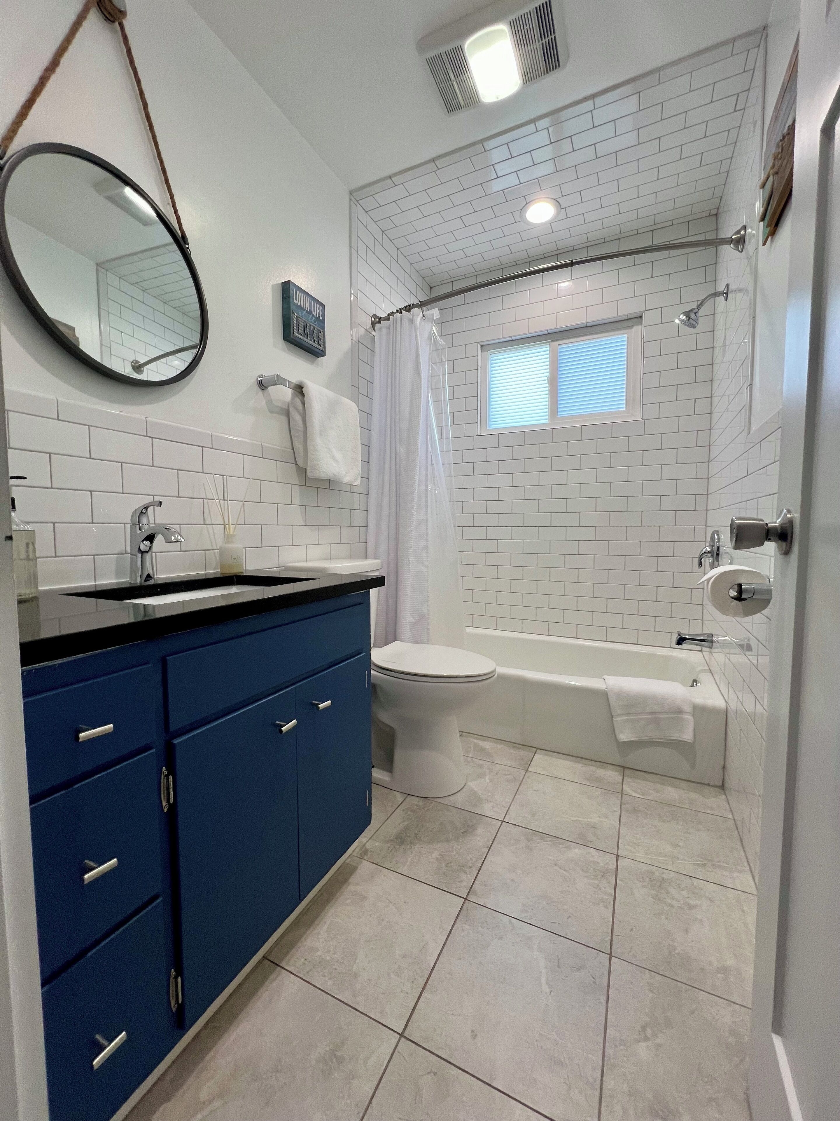 Um banheiro com paredes revestidas de azulejos brancos, um espelho com bancada azul, vaso sanitário, banheira e cortina de chuveiro.
