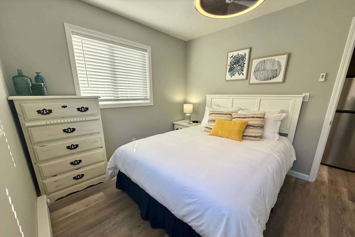 Une chambre à coucher comprenant un lit, une commode, une fenêtre, des œuvres d’art et un réfrigérateur.