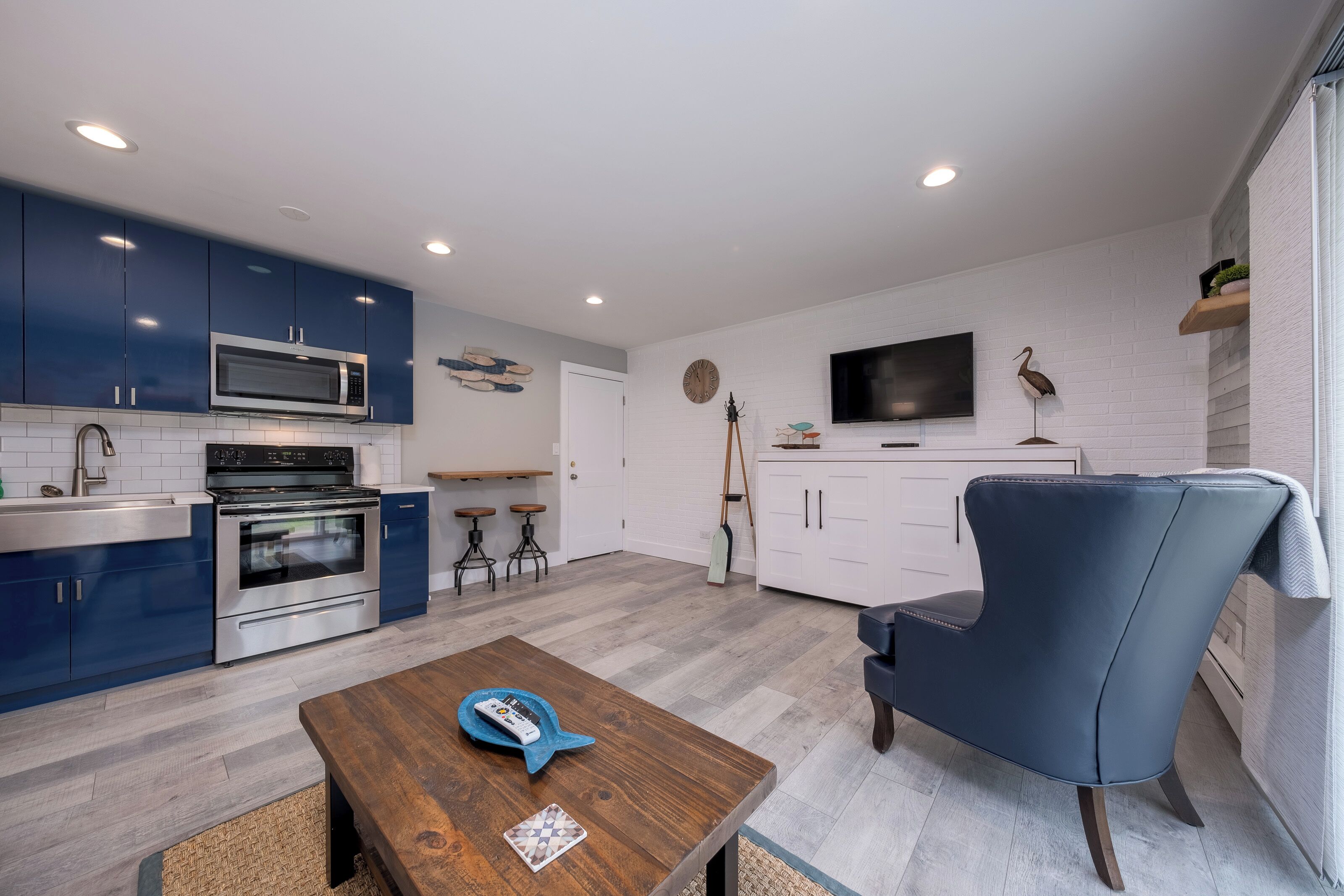 Cocina moderna con armarios azules, microondas y horno. Hay una mesa de madera con un plato de pescado y un control remoto. Un sillón azul está al lado de un mueble blanco.