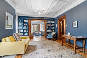 A room with a yellow sofa, a patterned rug, a wooden table, and a bookshelf filled with books and decorative items. - Spacious Historic 3-Bedroom Apt on Quiet Tree-Lined Street Near Peace Bridge (Buffalo)