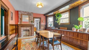 A dining area with a wooden table and chairs, a brick fireplace, and a window with plants.