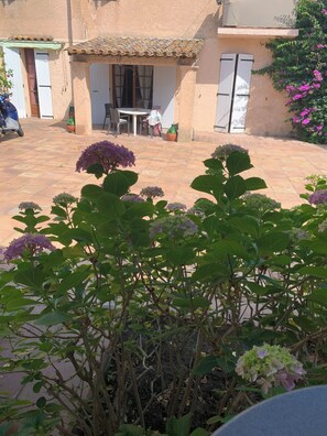 Um pátio com mesa e cadeiras, cercado por plantas e um edifício com telhado de azulejos.