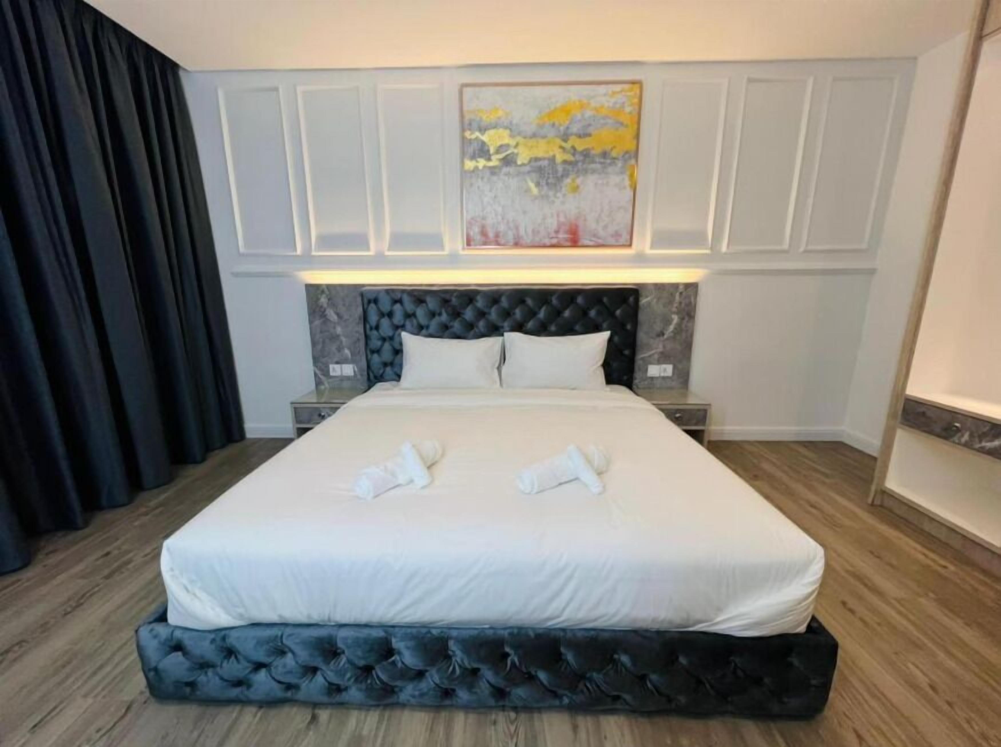 A hotel room with a large bed, a headboard with a tufted design, a painting above the bed, and a dark curtain on the left.