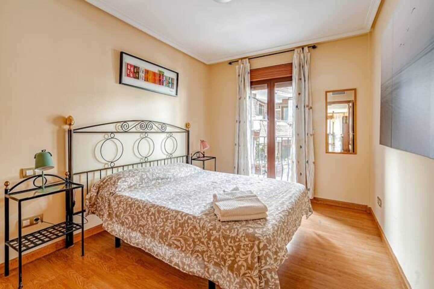 Un dormitorio con cama, mesita de noche, lámpara, ventana con cortinas y un cuadro en la pared.