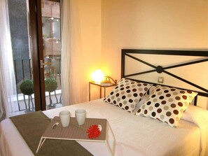 Habitació d'hotel amb un llit, una taula de nit amb una làmpada, una safata amb tasses i una flor, i vista al balcó exterior.