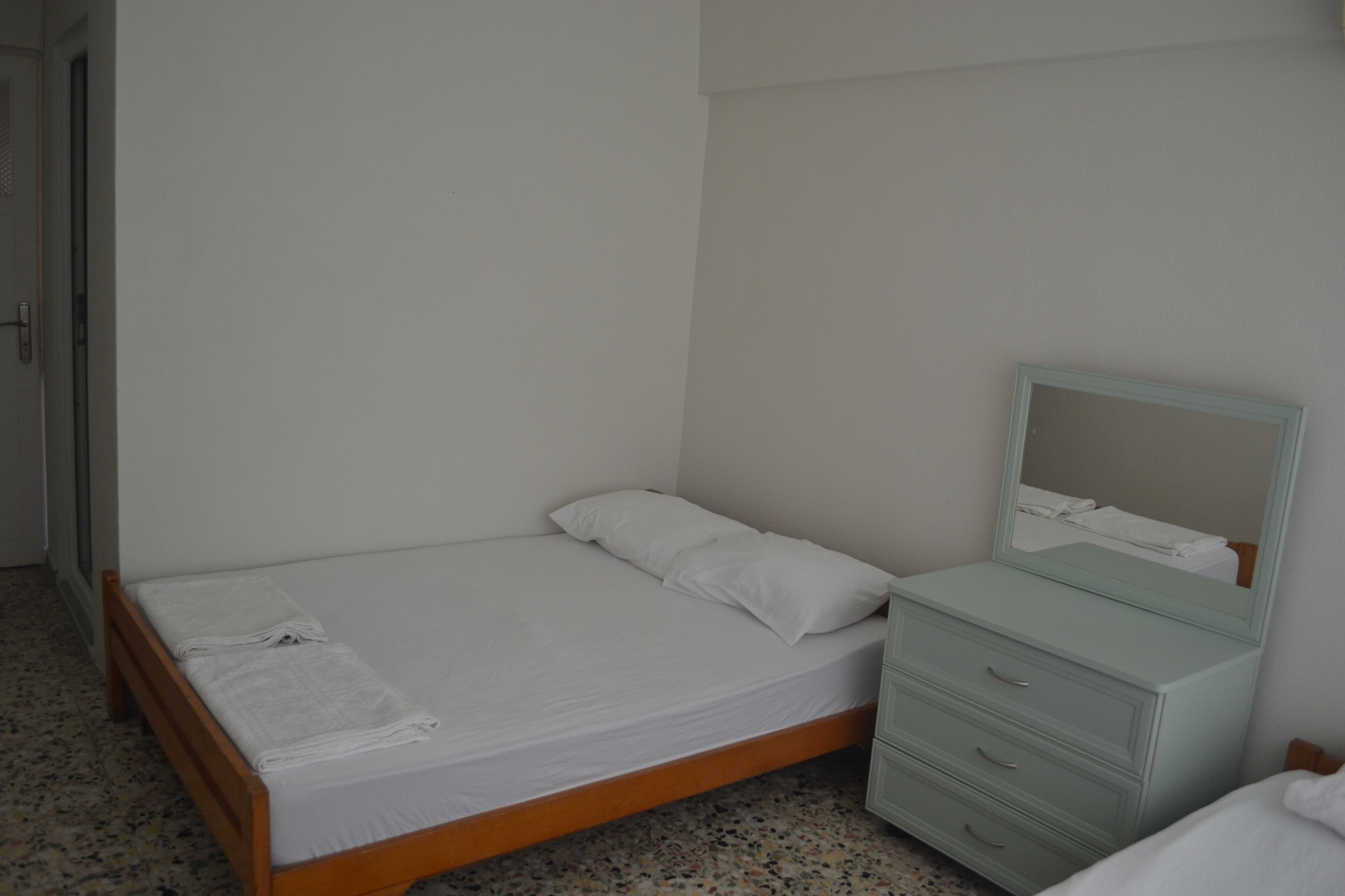Una habitación pequeña con cama individual, estructura de madera, colchón y almohada blancos, una mesita de noche con cajones y un espejo sobre la mesa.