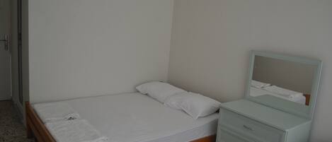 Una habitación pequeña con cama individual, estructura de madera, colchón y almohada blancos, una mesita de noche con cajones y un espejo sobre la mesa.