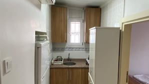 A narrow kitchen with wooden cabinets, a refrigerator, and a window. - YAZICI (Edremit)