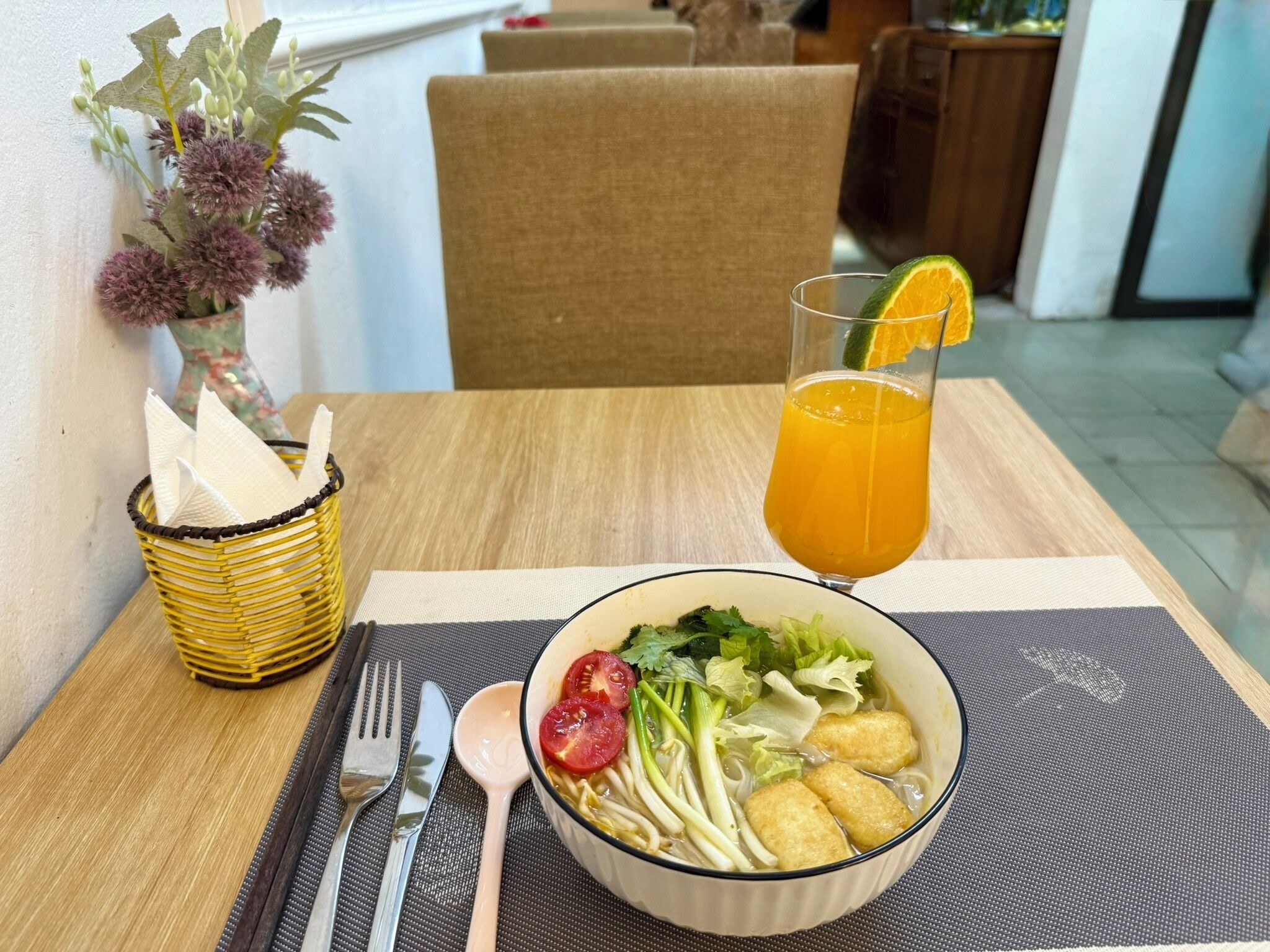 A dining table with a bowl of soup, a glass of orange juice, a napkin holder, and a fork and spoon set.