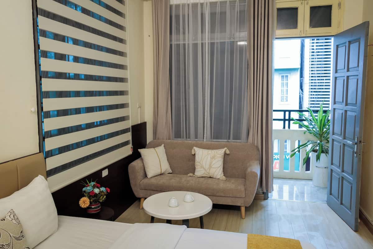 A hotel room with a sofa, a small round table, a bed, a window with sheer curtains, and a view of a balcony with a plant.