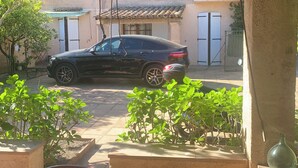 A covered outdoor area with a car parked under a wooden roof, surrounded by potted plants and a tiled floor.