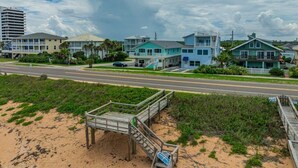 Ein Strandgrundstück mit einer Holzstege, der zum Strand führt, einer Straße im Vordergrund und mehreren Gebäuden im Hintergrund.