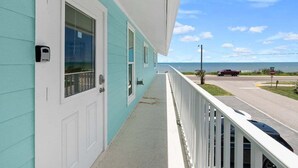 A turquoise building with a white door and windows, a balcony with a white railing, and a view of the road and ocean. - Fall Deals + Snowbird Rates | Ocean Views + Beach Getaway (Flagler Beach)