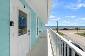 Ein türkisfarbenes Gebäude mit weißer Tür und Fenstern, einem Balkon mit weißer Brüstung und Blick auf die Straße und das Meer.