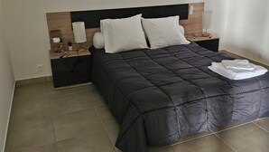A bedroom with a black bedspread, two white pillows, a wooden headboard, a nightstand with a lamp, and a small potted plant. - Lower villa F3 (Alata)