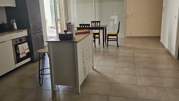 A modern kitchen with a central island, a dining area with a table and chairs, and a large sliding glass door leading to a balcony. - Lower villa F3 (Alata)