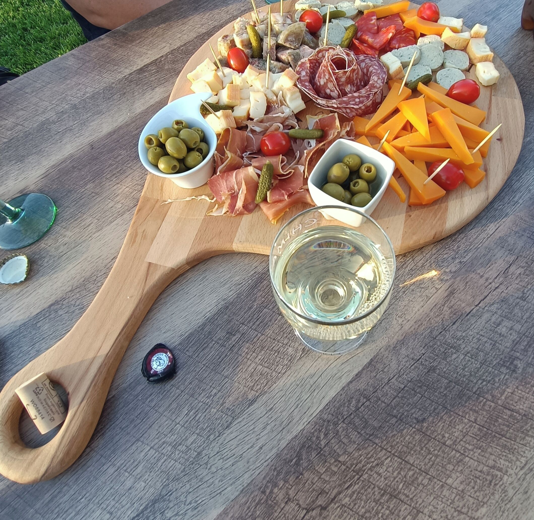 Ein Holzbrett mit verschiedenen Käsesorten, Fleisch und Oliven, begleitet von einem Glas Wasser und einer Bierkappe.