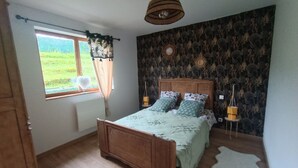 A bedroom with a wooden bed, a window with curtains, a patterned wall, and a hanging light fixture. - Le chalet du Lilas (Liessies)