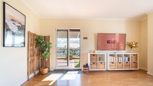 A modern hotel lobby with a wooden floor, a sliding glass door leading to a balcony, a flat-screen TV, and a bookshelf with wicker baskets.