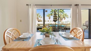 Un coin repas avec une table en bois dressée pour quatre personnes, une vue sur une piscine et des palmiers, et une porte-fenêtre coulissante.