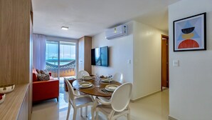Un comedor moderno con una mesa de madera dispuesta para cuatro, sillas blancas y un sofá rojo con cojines estampados. Un televisor de pantalla plana está montado en la pared sobre la mesa.