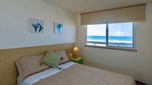 Un dormitorio con cama, mesita de noche con lámpara, dos cuadros en la pared y ventana con vista al océano.