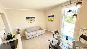 A modern living room with a sofa, a glass dining table, and two chairs. There are two paintings on the walls, a kitchen area with a stove, and a window with a view of greenery. - Villa Ipnotica - Panoramic Views Plus Exquisite Pool - Sleeps 7 - Very Stylish (Spoleto)