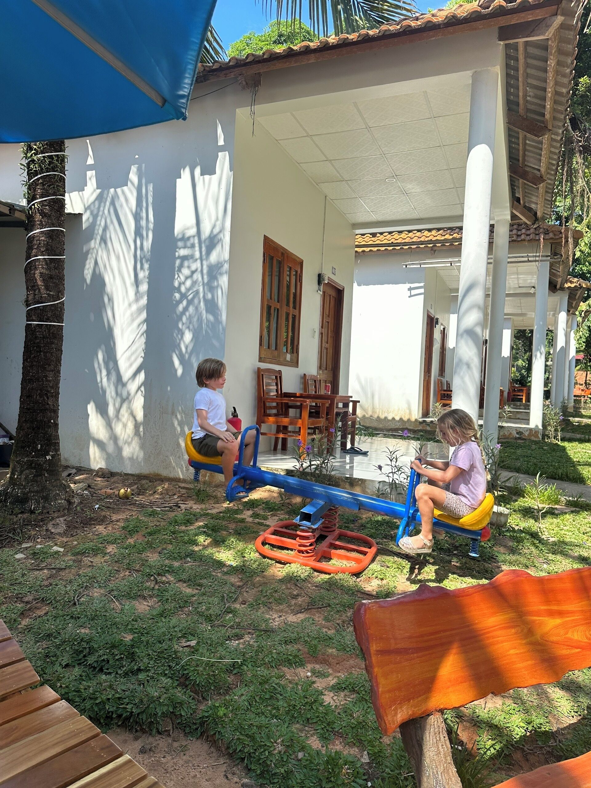 Two children playing on seesaws in a covered outdoor area with a white wall and wooden furniture.