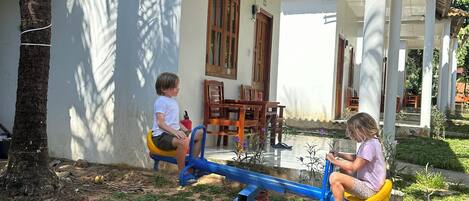 Two children playing on seesaws in a covered outdoor area with a white wall and wooden furniture.
