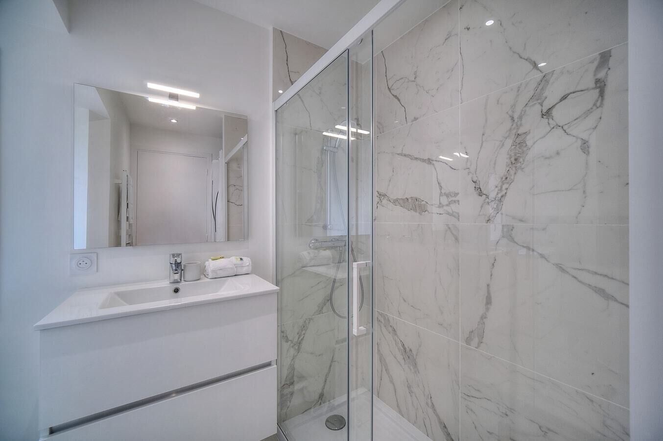 Een moderne badkamer met een douchecabine van glas, een witte wastafel met spiegel en marmeren tegels.