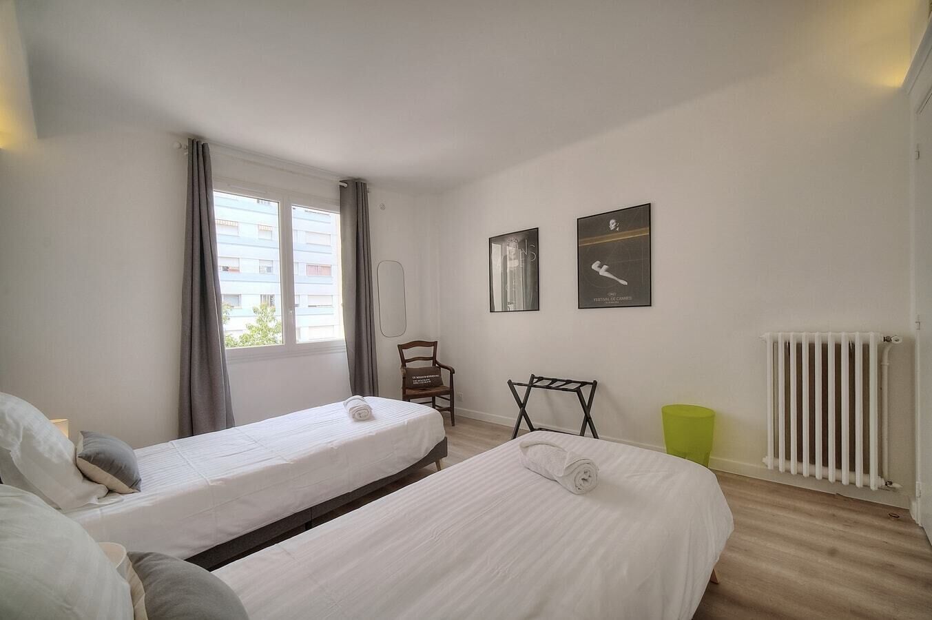 Een hotelkamer met twee bedden, een radiator, een raam met gordijnen en twee foto's in lijsten aan de muur.