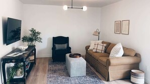 Ein Wohnzimmer mit schwarzem Sessel, beiger Couch, Couchtisch, Fernseher an der Wand und einer Pflanze.
